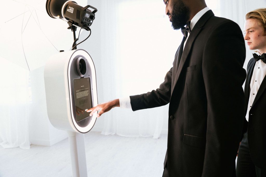 Two men in tuxedos stand next to a wedding photo booth.